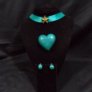May include: A teal green heart-shaped pendant necklace with a gold star charm on a teal green ribbon choker. Matching teal green round stud earrings are also included.