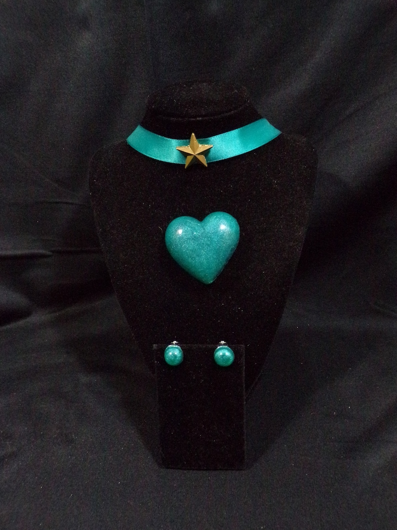 May include: A teal green heart-shaped pendant necklace with a gold star charm on a teal green ribbon choker. Matching teal green round stud earrings are also included.