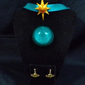Pode incluir: Um colar choker azul turquesa com um pingente de estrela dourada e um pingente de orbe azul turquesa combinando. O colar &eacute; exibido em um suporte de veludo preto com um par de brincos de planeta dourados.