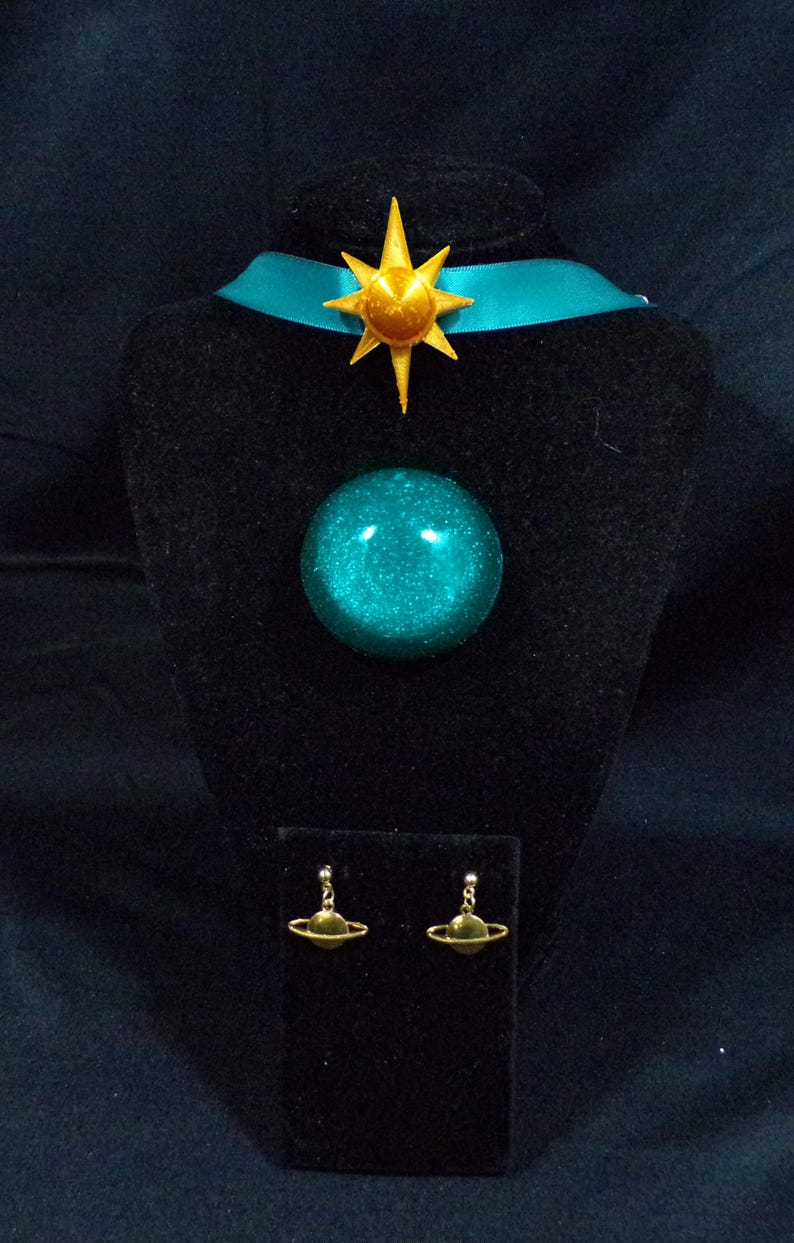 Pode incluir: Um colar choker azul turquesa com um pingente de estrela dourada e um pingente de orbe azul turquesa combinando. O colar &eacute; exibido em um suporte de veludo preto com um par de brincos de planeta dourados.