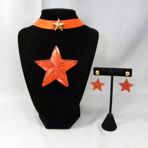 May include: A red glitter star necklace and earrings set. The necklace features a red glitter star pendant and a red satin ribbon choker with a gold star charm. The earrings are dangle earrings with red glitter star charms.
