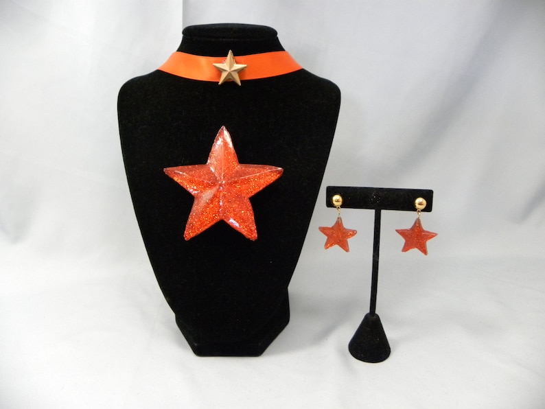May include: A red glitter star necklace and earrings set. The necklace features a red glitter star pendant and a red satin ribbon choker with a gold star charm. The earrings are dangle earrings with red glitter star charms.