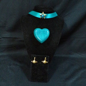 May include: Teal blue choker necklace with a gold star charm, a teal blue heart-shaped pendant, and gold planet earrings. The jewelry is displayed on a black velvet stand.