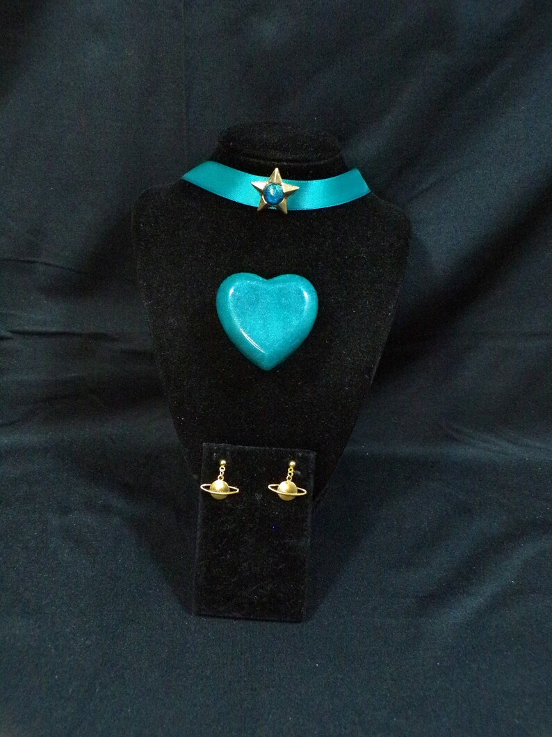 May include: Teal blue choker necklace with a gold star charm, a teal blue heart-shaped pendant, and gold planet earrings. The jewelry is displayed on a black velvet stand.