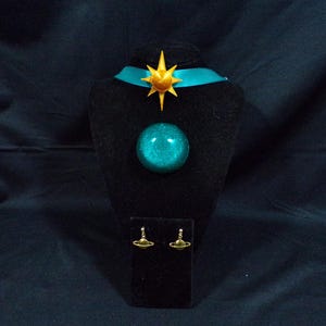 Pode incluir: Um colar choker azul turquesa com um pingente de estrela dourada e um pingente de orbe azul turquesa correspondente. O colar &eacute; exibido em um suporte de veludo preto com um par de brincos dourados com um pequeno pingente de orbe azul turquesa.