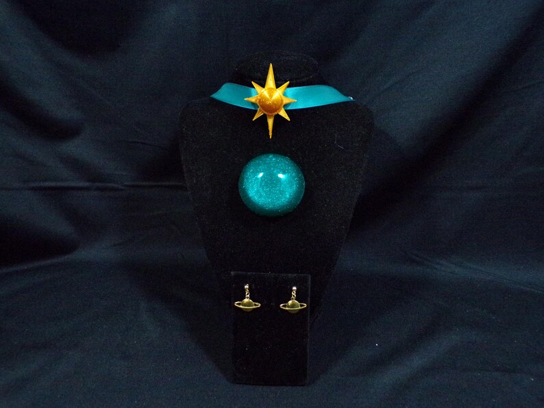 Pode incluir: Um colar choker azul turquesa com um pingente de estrela dourada e um pingente de orbe azul turquesa correspondente. O colar &eacute; exibido em um suporte de veludo preto com um par de brincos dourados com um pequeno pingente de orbe azul turquesa.