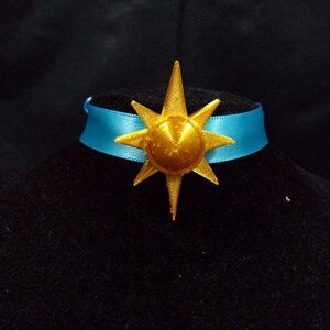 Pode incluir: Um pingente de estrela dourada com um colar de fita azul. A estrela tem um centro elevado e uma superf&iacute;cie texturizada.