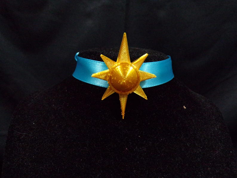 Pode incluir: Um pingente de estrela dourada com um colar de fita azul. A estrela tem um centro elevado e uma superf&iacute;cie texturizada.