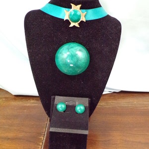 May include: A teal green and gold statement necklace with matching earrings. The necklace features a large, round pendant and a starburst design with a teal green stone. The earrings are small and round.