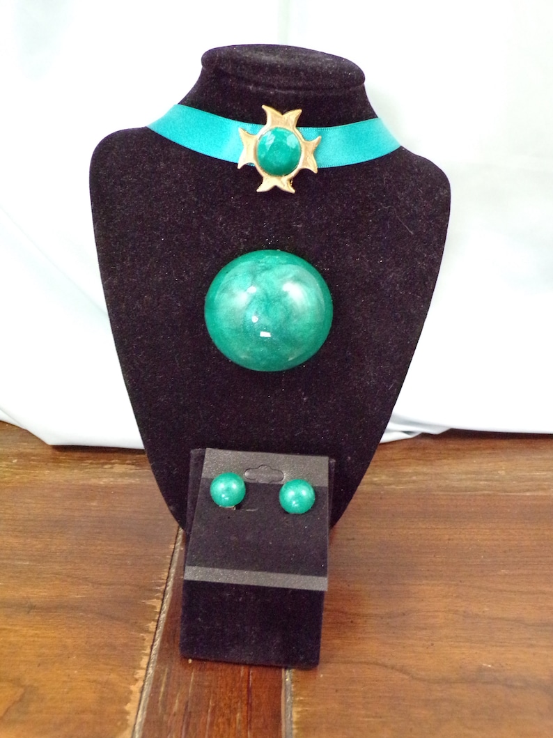 May include: A teal green and gold statement necklace with matching earrings. The necklace features a large, round pendant and a starburst design with a teal green stone. The earrings are small and round.