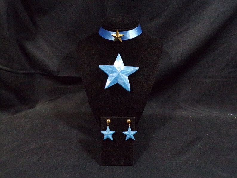 May include: A blue star-shaped necklace, earrings, and choker set. The necklace and earrings have a light blue, iridescent finish. The choker is made of blue ribbon with a gold star charm.