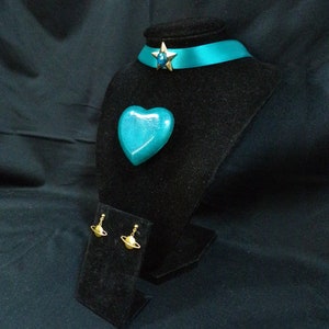 May include: Teal blue heart-shaped pendant, a teal blue ribbon choker with a gold star charm, and gold planet earrings on a black velvet display stand.