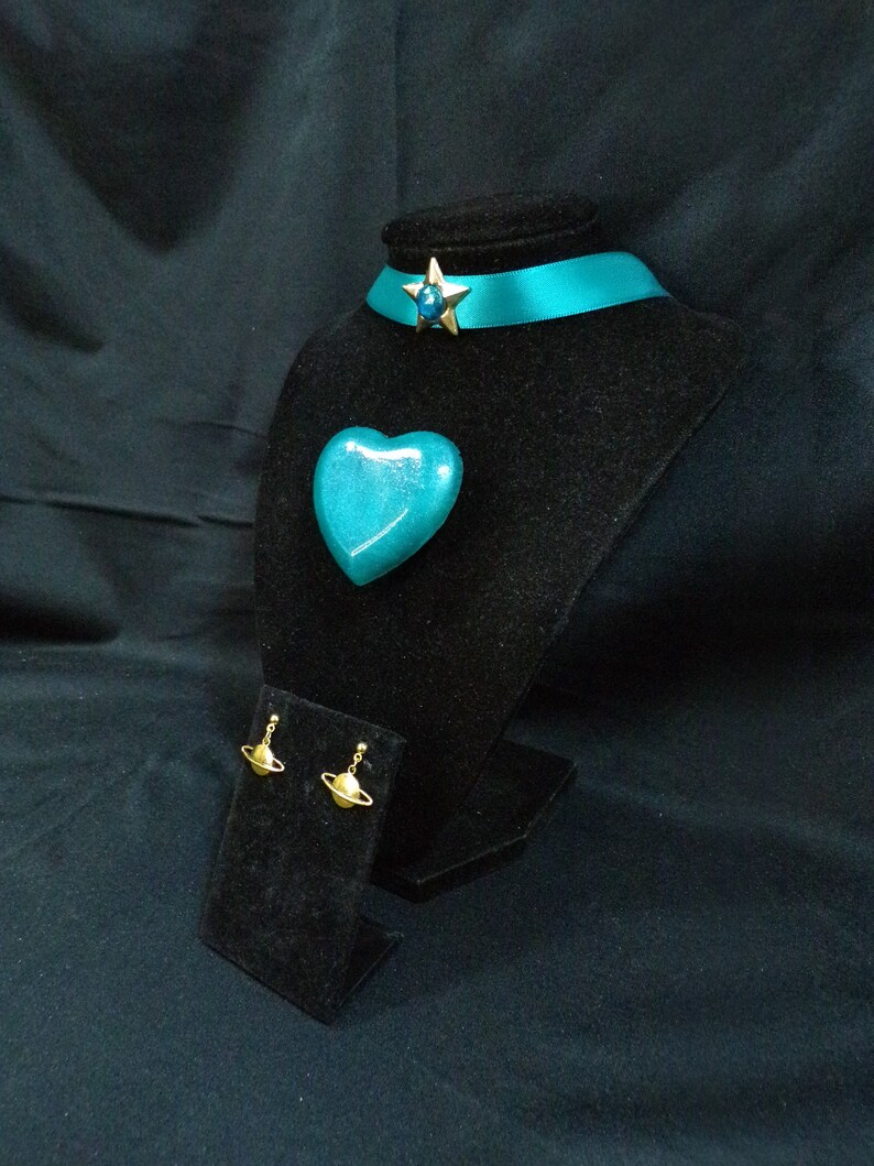 May include: Teal blue heart-shaped pendant, a teal blue ribbon choker with a gold star charm, and gold planet earrings on a black velvet display stand.