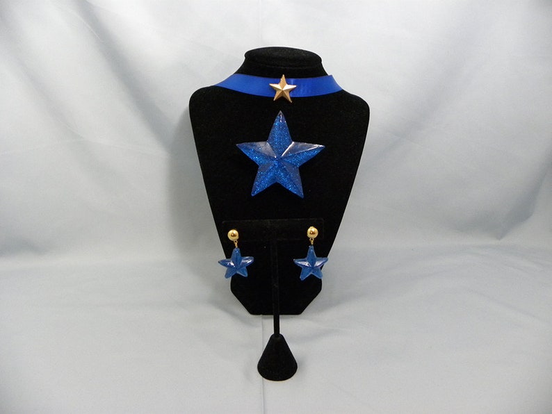 May include: A blue glitter star necklace and earrings set. The necklace features a blue glitter star pendant and a blue ribbon choker with a gold star accent. The earrings are blue glitter stars with gold posts.