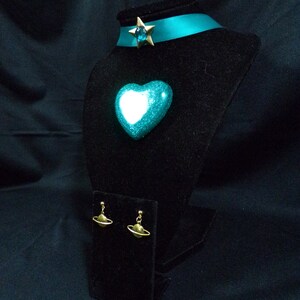 May include: A teal blue heart-shaped pendant with a gold star charm and a teal blue ribbon choker. The pendant and choker are displayed on a black velvet stand. The earrings are gold and feature a small sphere with a ring around it.
