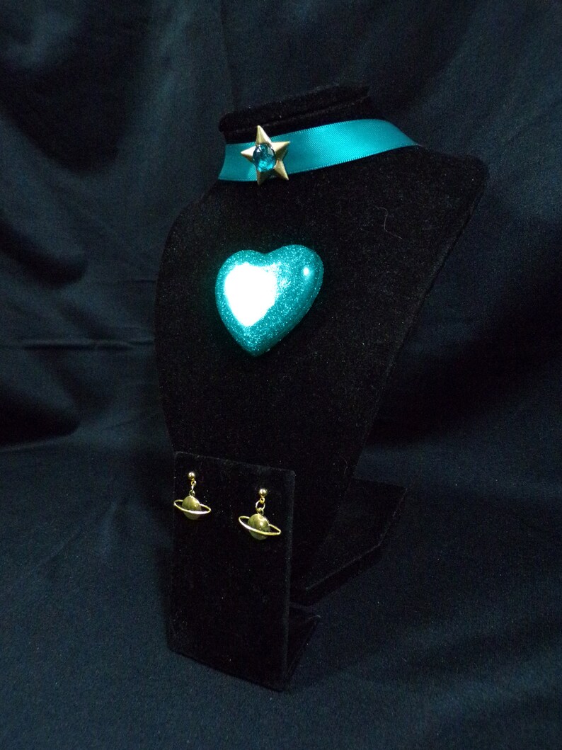 May include: A teal blue heart-shaped pendant with a gold star charm and a teal blue ribbon choker. The pendant and choker are displayed on a black velvet stand. The earrings are gold and feature a small sphere with a ring around it.