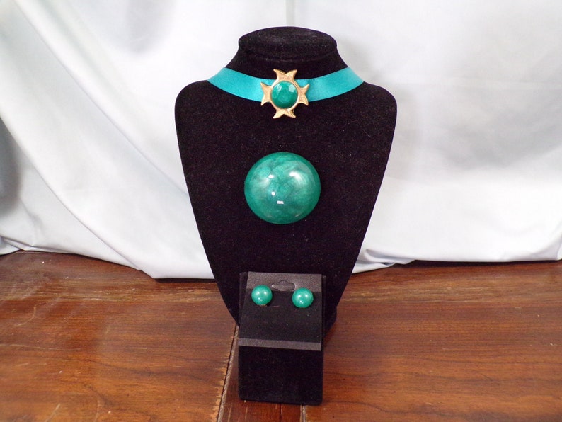 May include: A turquoise and gold jewelry set with a large round pendant, a smaller round brooch, and matching earrings. The set is displayed on a black velvet stand.