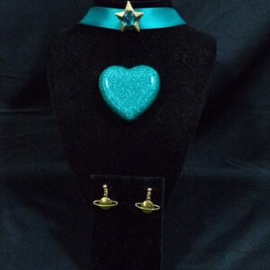 May include: A turquoise glitter heart pendant necklace with a gold star charm and matching turquoise glitter earrings with gold planet charms.