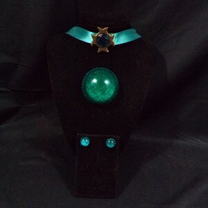 May include: A teal blue choker necklace with a gold star charm and a large teal blue gem pendant. Matching teal blue gem earrings are displayed on a black velvet stand.