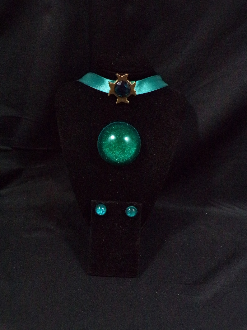 May include: A teal blue choker necklace with a gold star charm and a large teal blue gem pendant. Matching teal blue gem earrings are displayed on a black velvet stand.