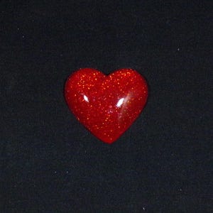 May include: A red heart-shaped object with a glittery surface. The heart is glossy and reflects light, set against a dark background. The shape is a classic symbol of love and affection.