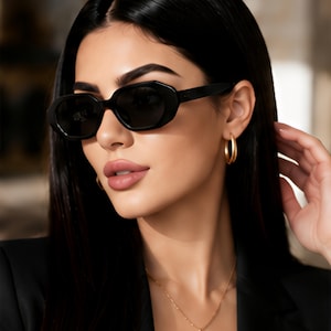 May include: Black sunglasses with thick, geometric frames and dark lenses. The eyewear features a bold, angular design with wide temples. The accessory is paired with gold hoop earrings and a delicate gold chain necklace with a small circular pendant.