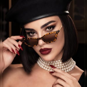 May include: A black beret with a leather trim, cat-eye sunglasses featuring a brown leopard print frame and dark lenses, and a multi-strand white pearl choker necklace. The accessories are worn with dark hair, red nail polish, and dark lipstick.