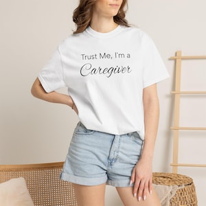 May include: A white short-sleeved t-shirt featuring the black printed text Trust Me, I'm a Caregiver. The shirt has a crew neckline and a standard fit, paired with light blue denim shorts that have a cuffed hem and visible stitching details.