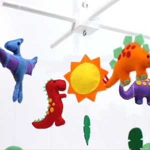 May include: A colorful felt dinosaur baby mobile with a yellow sun, green leaves, and purple, orange, red, and blue dinosaurs.