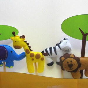 May include: Four felt animal puppets on sticks, including a blue elephant, yellow giraffe, black and white zebra, and brown lion. The puppets are set against a white background with green felt trees.