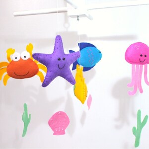 Könnte beinhalten: Ein farbenfrohes Filz-Babymobil mit einem lila Seestern, einem orangefarbenen Krebs, einem blauen Fisch, einer rosa Qualle und einer gelben Muschel. Das Mobile enthält außerdem zwei grüne Seegras-Dekorationen.