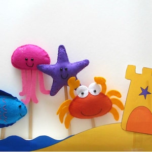 May include: A set of felt sea creature puppets, including a blue fish, a pink jellyfish, a purple starfish, and an orange crab. The puppets are on sticks and there is a yellow felt sandcastle in the background.