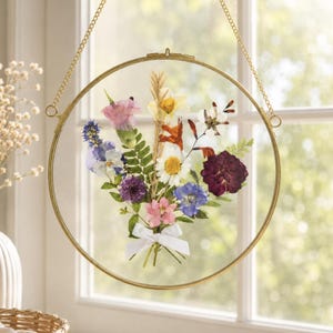 May include: A circular gold-toned metal frame holds a pressed flower arrangement between two glass panes. The bouquet features purple, pink, white, and yellow dried flowers with green ferns, tied at the base with a small white ribbon.