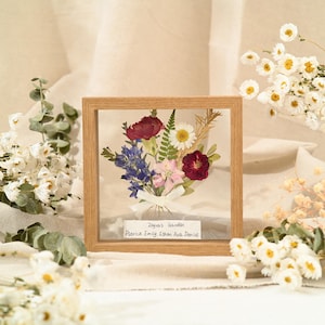 Op de afbeelding: Een vierkante lijst van licht hout met geperste bloemen in rood, blauw, roze en wit, versierd met een klein wit lintje. Een wit label onderaan bevat de tekst: Zayra's Garden, Patrick, Emily, Ethan, Ava, Denise.