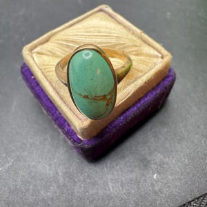 May include: A gold ring featuring a large, oval-shaped turquoise stone with visible brown matrix veining. The stone is set in a smooth gold bezel, and the band is a simple, polished gold metal. The ring is displayed on a small, square velvet box.