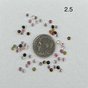 May include: A collection of small, round, faceted gemstones in shades of pink, brown, and clear, scattered around a silver-colored United States dime coin. The number 2.5 is printed in the upper right corner, indicating a size of 2.5 millimeters.