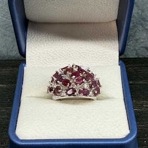 May include: A silver-toned ring featuring a cluster of numerous oval-cut, deep red gemstones arranged in a dome shape. The ring is presented inside a dark blue, hinged jewelry box with a cream-colored interior lining and a textured surface.