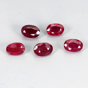 May include: Five oval-cut rubies with a deep red color and faceted surfaces. Each gemstone displays a polished finish with multiple triangular facets that reflect light, showcasing the vibrant red hue and clarity of the stones arranged in a group.