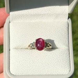 May include: A silver ring featuring a central oval-shaped cabochon-cut deep pink gemstone. The metal band displays a decorative openwork design on both sides of the stone. The ring is presented inside a light gray hinged jewelry box with a white interior.