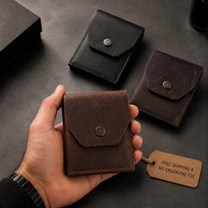May include: Three small leather wallets with a flap closure and a single metal snap button. One wallet is black, while two are dark brown. The wallets feature a rectangular shape with visible stitching along the edges and a smooth matte finish.