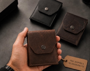 May include: Three small, rectangular leather wallets with a flap closure secured by a single metal snap button. One wallet is black, while two are dark brown with a textured finish. A tan paper tag is attached to one wallet with string.