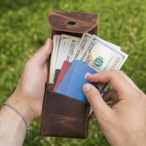Może przedstawiać: Brązowy skórzany portfel z zapięciem na zatrzask, zawierający kilka amerykańskich banknotów studolarowych oraz kilka pustych kart kredytowych w kolorze niebieskim, czerwonym i czarnym, trzymany w sposób otwarty, aby ukazać zawartość.