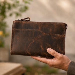 Può includere: Una pochette rettangolare in pelle marrone scuro con chiusura a zip superiore, linguetta in pelle abbinata e una tasca esterna a bustina con bordo tagliato in diagonale. La pelle presenta una finitura testurizzata e vissuta.