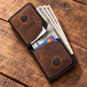 May include: A brown leather wallet with black trim and a black button closure on each side. The wallet is personalized with the name JAMES engraved on the front panel. It contains several US dollar bills and a blue and red payment card inside.