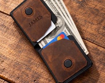 May include: A brown leather wallet with black trim and a black button closure on each side. The wallet is personalized with the name JAMES engraved on the front panel. It contains several US dollar bills and a blue and red payment card inside.