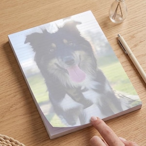 May include: A rectangular notepad featuring a printed image of a black, white, and tan dog with its tongue out. The notepad has horizontal lines for writing and is positioned on a wooden surface next to a pen and a small glass vase with lavender.