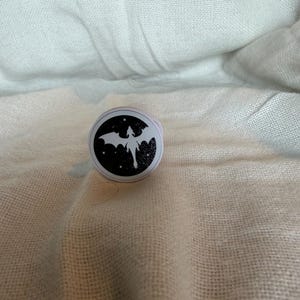 Könnte beinhalten: Ein runder Handyhalter mit schwarzem Hintergrund und einer weißen Silhouette eines fliegenden Drachen, umgeben von kleinen weißen Sternen. Das Zubehör hat einen weißen Außenrand und ein zusammenklappbares Design für einen sicheren Halt.