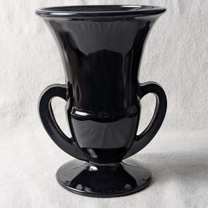 May include: A glossy black glass vase featuring a wide, flared rim, a tapered body, and two symmetrical curved handles on the sides. The vessel stands on a circular pedestal base, showcasing a smooth, reflective surface throughout the entire piece.