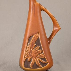May include: A tall, tapered ceramic pitcher in a matte terracotta orange finish. The body features a recessed, dark brown panel with a raised, molded floral design of a flower and wheat stalk. It has a curved handle and a dark base.