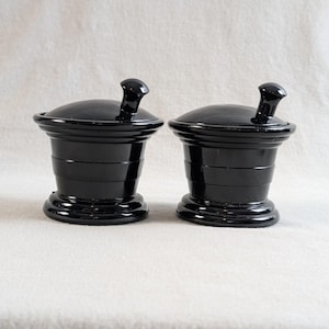 May include: Two matching black glass jars with lids, each featuring a small knob handle on top. The jars have a wide, flared rim, a tapered body with horizontal ridges, and a circular base, presenting a smooth, glossy finish throughout.